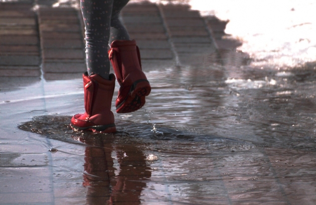 The lower half of a child wearing red rainboots and splashing in a puddle on a brick street.